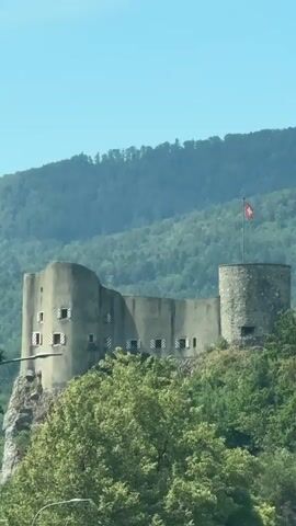 The video shows a fortress on a hill with trees in the foreground and a mountain range in the background. The fortress has stone walls and a round tower with a Swiss flag on top. There are windows on the walls of the fortress. The sky is clear and blue.