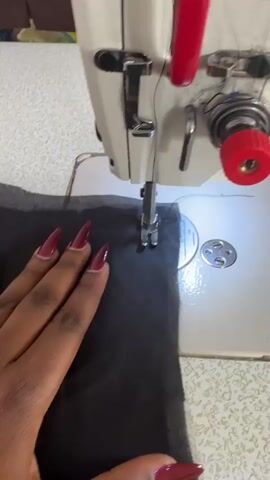 A close-up shot captures a person's hand operating an industrial sewing machine on a white work surface in Dushepe, Nigeria. The hand, likely female, features long, dark red manicured nails, guiding a piece of dark, sheer fabric under the machine's silver presser foot. The sewing machine, primarily white with red accents on its handles and tension knob, is actively stitching the fabric. Visible machine components include the needle, presser foot, needle plate with circular markings, and part of the thread tension assembly. The scene is brightly lit, suggesting an indoor workshop or studio setting during the day or under strong artificial light. The activity depicts a focused and precise sewing task.