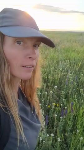 A woman wearing a gray baseball cap and a dark gray t-shirt is walking through a field of tall green grass with wildflowers. The sun is setting, casting a warm glow over the landscape. The woman is looking towards the horizon as she walks. The video is taken from a first-person perspective.