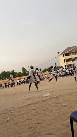 The video shows a volleyball game being played outdoors on a sand court. A group of people wearing white shorts and t-shirts, along with matching hats, are divided into teams and are actively playing the game. The volleyball net is stretched across the middle of the court, and players can be seen jumping, hitting, and setting the ball.  In the background, numerous individuals, also dressed in similar white attire, are seated on black chairs, observing the game. There are also some buildings and trees visible in the background. Debris like white paper litters the sandy surface of the court.