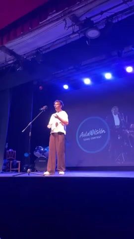 A young male with short, dark hair, wearing glasses, a white t-shirt, and loose brown pants, stands center stage, singing into a microphone. He is holding his hands clasped together in front of his stomach. Behind him, a large screen displays the words "AulaVision SONG CONTEST" within a blue circle and heart graphic, with a silhouette of a band playing on the right side. The stage is lit with blue spotlights. To the left of the singer, a drum set and a chair are visible. The overall environment appears to be an indoor performance venue.
