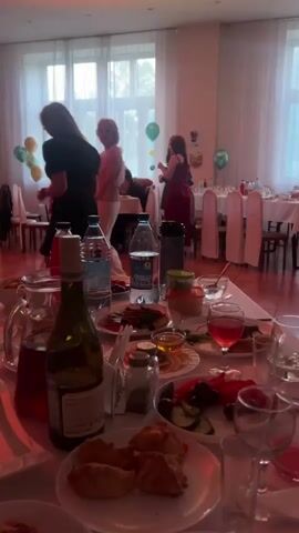 The video shows a gathering of people in a room that appears to be a banquet hall or restaurant. A long table laden with food and drinks is in the foreground, with several place settings and wine glasses visible. In the background, people are mingling and socializing. Some are standing and talking, while others appear to be dancing or moving around the room. The lighting is dim with some colored lights adding to the festive atmosphere. Balloons are visible in the background, suggesting a celebration. The overall scene depicts a lively social event.
