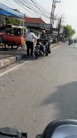 The video shows a street scene with several people and vehicles. In the foreground, a portion of a motorcycle's handlebars and dashboard is visible, suggesting the video is shot from the perspective of a motorcyclist. On the left side of the street, there is a stall with a large red tank and an engine on a cart. Two individuals are interacting near this stall. One, wearing a light-colored jacket and dark pants, appears to be handing something to another person who is sitting on a black motorcycle. This person on the motorcycle is wearing a helmet and a dark jacket. The street itself is paved and appears to be a public road, with a curb on the left side. Further down the street, there are buildings lining the right side, and several other motorcycles and possibly a car are visible in the distance. Overhead, power lines and utility poles are present. The overall environment suggests a daytime scene in a moderately populated area, possibly a market or a street with businesses. The interaction between the two individuals near the stall seems to be a transaction or exchange of goods.
