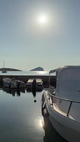 The video shows a harbor scene with several boats docked in calm water. The sun is high in the sky, casting a bright glare and reflecting on the water's surface. In the foreground, a white boat is partially visible on the right side of the frame, covered with a white tarp. Behind it, three smaller boats are moored in a line. The water is still and reflects the sky and the boats. In the distance, a small island with a dark, triangular shape can be seen. The sky is clear and pale blue.
