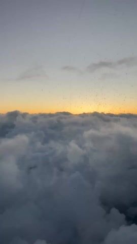 The video shows an aerial view from an airplane window. Below, a thick layer of clouds stretches to the horizon, with a band of orange and yellow light indicating the sun setting or rising. The airplane's engine is visible in the lower right corner of the frame, and part of the wing with the "Ryanair" logo is also visible. The window has some water droplets or condensation on it. The sky above the clouds is a mix of light gray and hints of orange from the sun.