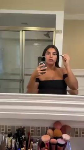 The video shows a young woman standing in a bathroom, facing a mirror. She is holding a smartphone in one hand and using an eyeliner brush in the other to apply makeup to her upper eyelid. She is wearing a black, off-the-shoulder top. The bathroom features a tiled shower with a glass door, a white bathtub, and a variety of makeup brushes and cosmetics arranged on a shelf beneath the mirror. The lighting is bright, and the woman's reflection is clear in the mirror.
