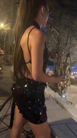 A woman is playing a saxophone. She is wearing a black sequined dress and her hair is in a ponytail. She is standing on a stone ledge with trees and lights in the background. There are tables and chairs in the background, suggesting an outdoor seating area.