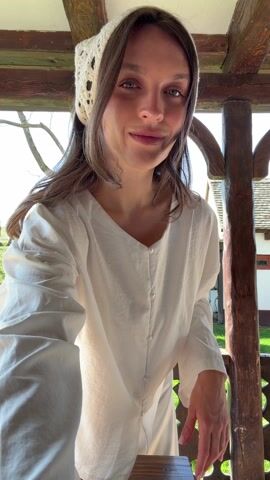 A young woman stands on a porch wearing a white blouse, white skirt, and a crocheted head scarf. The porch is made of dark wood and has a decorative railing. Behind her, a green lawn slopes upward towards a small building with a dark roof. A bare tree stands to the left of the building, and the sky is partly cloudy. She smiles at the camera, then gently adjusts her head scarf before looking off to the side and closing her eyes for a moment.