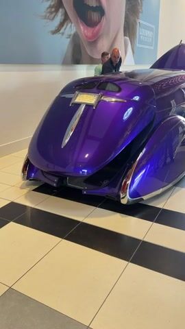 A medium-shot video shows the rear of a custom purple car with a sleek, futuristic design. The car is on display in what appears to be a museum, with a black and white checkered tile floor and a large poster on the wall behind it. Two men are standing near the rear of the car, looking at it. The poster on the wall features a close-up of a person's open mouth, with the text "LOUWWAN MUSEUM" visible. The car's design is characterized by its rounded curves, chrome accents, and a prominent fin-like structure at the back. The lighting is bright, highlighting the glossy finish of the car.