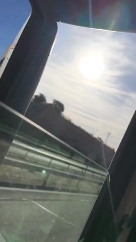 The video shows a view from inside a car traveling on a highway on a sunny day. The camera is positioned near an open car window, offering a partial view of the road and landscape outside. The sun is bright and high in the sky, with some clouds visible. The highway has multiple lanes and a median barrier. A car can be seen driving in the distance. The surrounding landscape includes grassy hills and an overpass. The inside of the car is dark, with the car door frame visible.