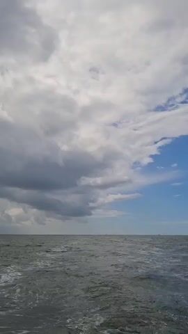 The video shows a view of the ocean from a moving vessel. Initially, the sky is partly cloudy with a mix of gray and blue patches. The water is dark with small waves. The camera angle shifts up to capture more of the sky, which then reveals predominantly blue sky with scattered white clouds. The camera shifts back down, and the sky turns overwhelmingly dark gray with heavy clouds covering the view. The sea remains choppy. The vessel continues to move through the water.