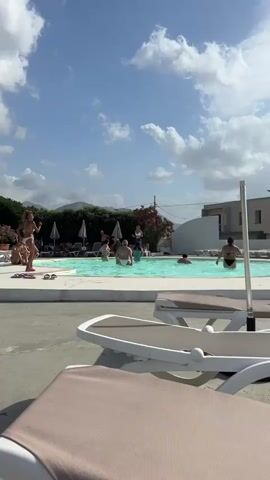 The video captures a sunny day at an outdoor swimming pool, likely during the daytime given the bright sky and shadows. The scene is set in Capaci, Italy, suggested by the provided context.

Several people are present. In the foreground, two empty lounge chairs with beige fabric are visible, implying a relaxed atmosphere where people might sunbathe. In the middle ground, a group of people are enjoying the pool. A woman in a white bikini is standing at the edge of the pool, appearing to be dancing or posing. Other individuals are in the water, some wading, others swimming. In the background, more people are seen relaxing on lounge chairs under umbrellas, suggesting a resort or recreational area. A lush green hedge and some flowering plants add to the outdoor setting, and in the distance, hills or mountains are visible under a bright blue sky dotted with fluffy white clouds.

The overall mood appears to be cheerful and leisurely, consistent with a vacation or a day spent at a pool. The weather seems pleasant and warm, suitable for swimming and outdoor activities. There is no visible text in the video.
