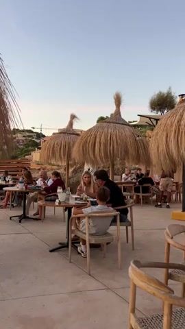 The video pans across an outdoor dining area set up on a paved surface, likely a patio or promenade, with a natural, rocky incline visible in the background. Several tables are occupied by groups of people, including families with children. The tables are topped with food and drinks, and some have umbrellas made of straw for shade. The atmosphere appears relaxed and casual, with individuals engaged in conversation and enjoying their meals. The lighting suggests it is either late afternoon or early evening, with a soft, warm glow. The overall setting evokes a sense of leisure and dining in a scenic, possibly coastal, environment.