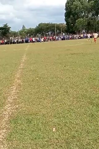 A video shows a soccer game in progress. The camera is initially focused on the field with a few players running downfield, with a crowd of spectators lining the perimeter. The camera suddenly moves upward as someone wearing a white shirt blocks part of the view.  The camera settles back down showing the soccer field again.
