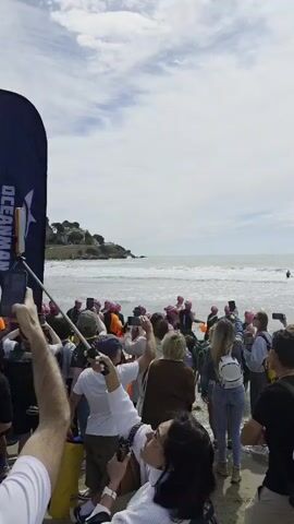 A large group of people are on a beach, many are entering the ocean, and the crowd is cheering them on, using their phones to record the event. The swimmers are wearing swimming caps and wetsuits, and the beach is sandy with a few waves. In the distance, a landmass can be seen. The sky is cloudy. Many people are wearing backpacks. Some people are wearing bicycle helmets.
