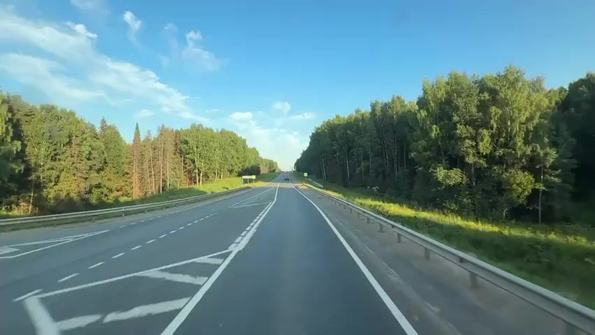 A first-person view from a vehicle driving on a highway through a forest. The road is paved with asphalt and has white lane markings. On both sides of the road, there are dense green forests. The sky is blue with a few white clouds. There are other vehicles visible on the road, including trucks. Traffic signs are present along the roadside. The sun is shining, casting shadows on the road and the surrounding vegetation. The overall impression is a scenic drive on a sunny day.