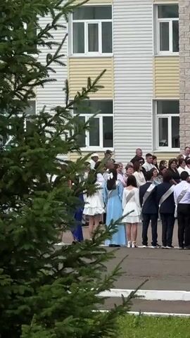 The video shows a group of people, likely students and adults, gathered outdoors in front of a building. In the foreground, a large evergreen tree with lush green needles partially obstructs the view. The building appears to be a multi-story structure with light-colored siding and prominent white-framed windows. Some windows have a pale yellow border. The overall impression is of a somewhat formal gathering.

The subjects are primarily young people dressed in what appear to be graduation or formal attire. Several young women are wearing white dresses, with one in a long, light blue dress. Some of the young men are dressed in dark suits with sashes, suggesting a ceremonial occasion. Adults, likely teachers or parents, are also present, standing behind the students.

The activity taking place seems to be a ceremony or a gathering before or after one, possibly a graduation ceremony given the attire of the students. The atmosphere appears festive yet orderly, with people standing in groups and facing the building. The lighting suggests it is daytime, likely late morning or early afternoon, with natural light illuminating the scene. The weather appears to be pleasant, with the greenery indicating a time of year with active plant growth, possibly late spring or summer.

No visible text is present in the video. The location cues, such as the building's architecture and the evergreen trees, are consistent with a school or similar institutional setting in a temperate climate. The gathering of young people in formal wear indicates a significant life event, likely related to education.