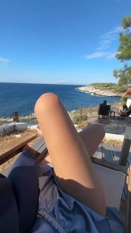 The video shows a first-person view of a person relaxing on a lounge chair outdoors, with a view of the ocean. The person's legs and knees are in the foreground, and they are wearing a light-colored silk robe or dress. The background features a calm, blue ocean with a coastline in the distance. There are a few other people visible in the background, sitting on chairs further away. The environment appears to be a scenic overlook or a terrace area near the sea, with some dry vegetation and a wooden deck. The sky is clear and blue, indicating good weather.