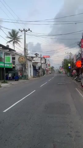 The video shows a street in a town or city. The street is paved and has white lines painted on it to indicate lanes. There are buildings on both sides of the street, some of which appear to be shops or businesses. There are also power lines and utility poles along the street. The sky is cloudy and the lighting suggests it might be early morning or late evening. There are a few vehicles visible, including a tuk-tuk parked on the left side of the street. There are also some signs on the buildings, including one that says "SINGER". The overall impression is of a quiet street, possibly before the day's activities have fully begun.
