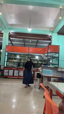 A woman wearing a black hijab and a blue skirt stands in what appears to be an indoor market or food stall area. She is looking to her right, with her left hand on her hip and her right hand holding a dark object, possibly a phone or a wallet. She is positioned in front of a display case filled with food items, which are arranged on shelves. The display case has an orange top and a glass front, allowing the food to be seen. Beneath the display case are cabinets with red and white labeling that reads "PRIME."

The setting is indoors with a high ceiling supported by light green beams. Overhead, there are several lights, including a prominent pendant lamp. The floor is tiled and appears to be light-colored. To the right of the woman, there is a table with several bottles of water and some other items. In the foreground, an orange chair is partially visible. The overall lighting suggests it could be daytime, but the indoor setting makes it difficult to determine the exact time of day or weather conditions. The atmosphere seems casual, possibly a place where people come to eat or purchase food. There are no clear indicators of the woman's emotion, though her pose might suggest she is waiting or attending to something.