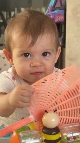 A close-up shot shows a baby sitting at a table playing with toys. The baby, with light brown hair and expressive eyes, wears a white shirt with a strawberry pattern. Their right hand is reaching towards a toy bee with black and yellow stripes, while their left hand holds a pink plastic sieve. The background is out of focus, with hints of a chair and some shelving. The overall lighting suggests an indoor environment, possibly during the day.