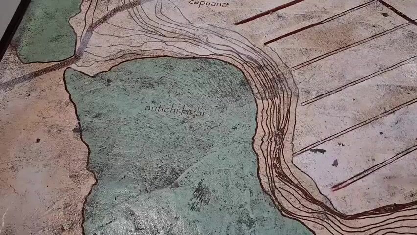 The video shows a close-up, panning view of a detailed map, likely etched or painted onto a surface. The map depicts an ancient city or settlement, featuring contour lines suggesting varied topography and bodies of water labeled as "antichi laghi" (ancient lakes). Several structures, roads, and entrances, including "Porta Capuana" and "Via Latina," are clearly marked. The map's color palette consists of muted earth tones with green accents for the water. The camera moves slowly, allowing for a detailed examination of the map's features. The lighting is somewhat diffuse, suggesting an indoor environment. There are no people visible in the video, and the focus remains solely on the map itself.