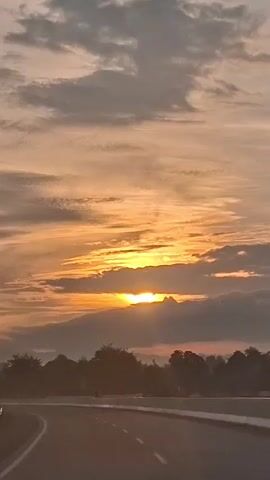 This is a vertical, medium-shot video of a sunset over a highway. The sky is filled with orange and gray clouds. The sun is visible through a gap in the clouds, casting rays of light. In the foreground, there is a highway with white lane lines. Trees and a low barrier are visible on the side of the road.