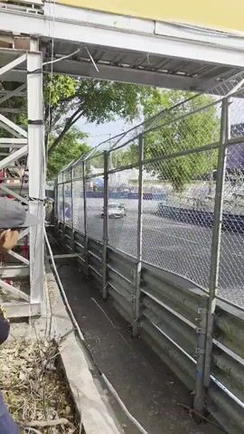 The video shows a race car speeding down a track. The camera is positioned behind a fence with a metal guardrail. Trees line the track, and the sky is visible through the leaves. The race car appears to be a white sports car. The video is short and jerky, suggesting it was filmed on a phone.