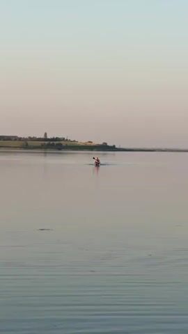 A kayaker is paddling on a calm body of water. The water is reflecting the clear sky, which is a soft pink and blue hue. In the distance, there is a strip of land with some trees and buildings. The kayaker is in the middle of the frame, and the boat is red. The sun is low in the sky, suggesting it is either sunrise or sunset. The overall mood is peaceful and serene.
