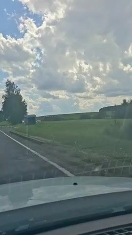 The video shows a view from inside a vehicle, looking out at a rural landscape. The sky is partly cloudy with sunlight breaking through. On the left side of the road, there's a bus stop with a shelter and a sign. Further down the road, there's a small building with a covered area and a trailer. A large tree is visible on the right side of the road, casting shadows. The road itself has a white line marking the lane. In the distance, there are fields and more trees, suggesting a countryside setting. The vehicle appears to be in motion, slowly moving forward.