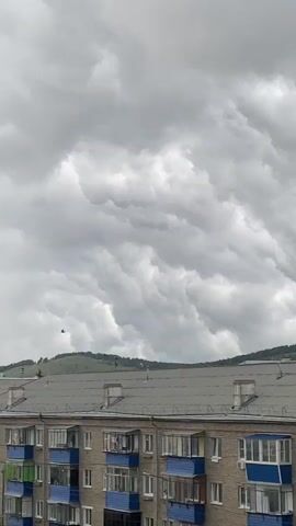 A video shows a building with a grey roof in the foreground. The sky is filled with dramatic, dark, and turbulent mammatus clouds. A small bird can be seen flying in the distance to the left. The overall scene suggests an impending storm.