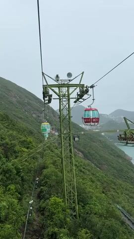 The video shows a cable car system in Hong Kong. The cable cars are brightly colored, with one being red and white and another being green and yellow. They are moving along cables supported by green metal towers. The landscape includes lush green mountainsides and a view of the ocean. Some buildings are visible in the distance across the water. The sky is overcast. A child can be heard speaking in the background.