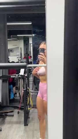 A woman wearing pink athletic shorts and a pink sports bra stands in a gym, holding a phone to take a selfie in a mirror. She is wearing headphones and has her hair tied back. Behind her, gym equipment and other people working out are visible.