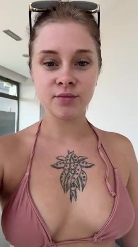 A young woman with blonde hair pulled back and sunglasses on her head, wearing a pink bikini top with a flower tattoo on her chest, looks into the camera. She briefly touches her hair and then looks away. The background appears to be an indoor hallway with a white wall and some windows.
