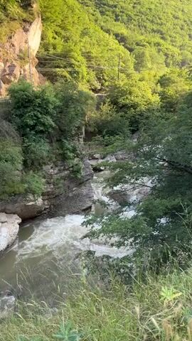 A wide shot captures a fast-flowing river cutting through a rocky gorge. Lush green trees and vegetation cover the steep slopes on either side of the river. The water is churning and white with rapids, indicating a strong current. The lighting suggests it is daytime, with sunlight filtering through the trees and illuminating parts of the scene. There are no people or animals visible in the video. The overall impression is of a natural, wild landscape.