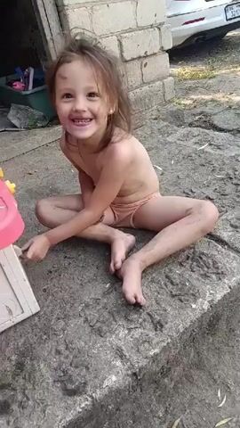 A young girl, appearing to be around 3-4 years old, sits cross-legged on a concrete surface. She is dirty, with mud smudges on her face, arms, and legs. She is wearing light-colored underwear. Her hair is messy and falls around her face. She has a wide, toothy smile and appears to be looking directly at the camera. Her eyes are bright and expressive, conveying a sense of joy and playfulness.

To her left, partially visible, is a pink and beige toy kitchen or playhouse. The girl's left hand is near this toy. The background shows a rough, unpainted wooden structure or shed on the left, and a brick wall on the right. Beyond the brick wall, a white car is partially visible. The ground around the girl is also concrete, with some fallen leaves and debris scattered on it. The scene appears to be outdoors, possibly in a yard or an area behind a building.

The lighting suggests it is daytime, likely with bright sunlight, though the directness of the light is hard to determine from the image alone. The overall impression is of a child enjoying some outdoor play, perhaps getting messy in the process. The emotion conveyed is one of uninhibited happiness and engagement. There is no visible text in the image.