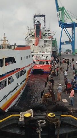 The video shows a port scene. In the foreground, a large, white and red ship is docked. The ship has multiple decks and windows, and some people can be seen inside. The ship is tethered to the dock by thick ropes. A large, black fender is also visible, attached to the dock. In the background, another ship, painted red and white, is docked alongside. A pier extends from the dock, lined with people. Some of these people appear to be passengers, while others may be port workers. Cargo cranes, painted green and blue, are visible in the distance, suggesting a busy shipping environment. The sky is overcast. The overall impression is that of a busy port with ships loading or unloading passengers and cargo.