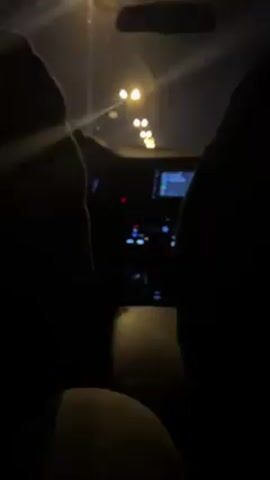 The video shows a view from the back seat of a car at night. Streetlights shine through the front windshield. The dashboard and center console are visible, but mostly dark. The backs of the front seats and the legs of the person in the back seat are also visible. The person seems to be moving slightly to the music playing.