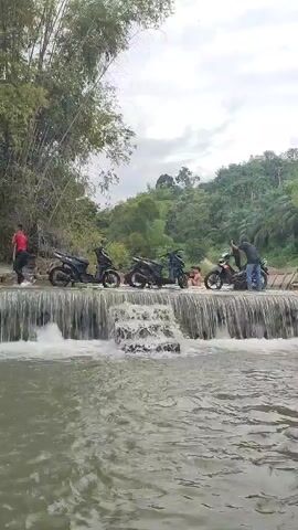 A video shows four motorcycles parked on a concrete dam with water flowing over it. Three people are visible near the motorcycles. One person wearing a red shirt stands on the left side of the dam. Two people are on the right side of the dam, with one person sitting on a motorcycle and another standing next to it. In the center of the dam, two people are sitting in the water. The background features lush green trees and vegetation, with a cloudy sky overhead. The water appears murky and brown.