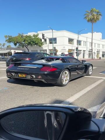 A black Porsche Carrera GT, featuring a California license plate "B063PO," is prominently displayed in the video. The vehicle is seen from a slightly elevated, rear-left perspective, suggesting the recording is made from inside another car, with a portion of a rearview mirror visible in the bottom-left corner of the frame.

The Porsche has a sleek, sporty design with large quad exhaust pipes, a visible engine cover with air intakes, and a subtle rear spoiler. The wheels are a light color, possibly silver or light gold, with a multi-spoke design. The interior appears to be light-colored, possibly white or silver.

The car is stopped or moving very slowly at what appears to be an intersection or crosswalk on a sunny day. As the video progresses, the car begins to accelerate, producing a distinct, high-pitched engine or exhaust sound.

The setting is an urban street lined with elegant, white two-story buildings, typical of a commercial or upscale retail district. Several tall palm trees are scattered along the street, adding to a warm-weather, possibly Californian, ambiance. The sky is clear and blue. Other vehicles, including a dark SUV and a white car, are visible in the background and foreground, indicating light traffic. The crosswalk is marked with white stripes on the asphalt.