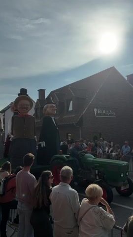 A parade is underway, featuring several large, costumed figures on a flatbed trailer pulled by a green tractor. A man drives the tractor, and at least three large figures are visible on the trailer. The figure closest to the viewer appears to be a woman wearing a dark skirt, a light-colored vest over a white shirt, and a dark hat. Next to her is a man in a dark suit and hat. To the far left, a third large figure, a man, is partially visible. A crowd of spectators stands behind a metal barrier, watching the parade. The scene takes place outdoors under a bright, sunny sky. The sun is high in the sky, causing some glare. A brick building with signage for "ELA Lighting" is visible in the background. The overall atmosphere suggests a festive or celebratory event.