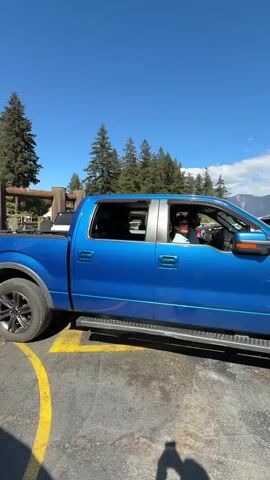 The video shows a blue Ford F-150 pickup truck parked in a ferry terminal lane on a clear, sunny day. The truck is an FX4 model with black wheels. The truck is parked at the Snug Cove Terminal on Bowen Island. The ferry ramp is visible in the background, raised and ready for vehicles to board. The ferry terminal has metal structures and signage. The surrounding environment includes trees and a clear blue sky. There are also some vehicles parked behind the pickup truck. There is a yellow warning sign that reads "ATTENTION: PLEASE REMAIN BEHIND YELLOW LINE UNTIL DIRECTED BY CREW".