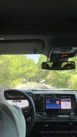 The video shows a first-person perspective from inside a car driving on a winding road surrounded by trees. The driver's hands are visible on the steering wheel. The car's dashboard displays a navigation system and a music player interface, showing Spotify and a song called "Agua Sana". In the rearview mirror, a person's face is partially visible. Another vehicle is seen ahead on the road. The sun is shining, indicating it is daytime.