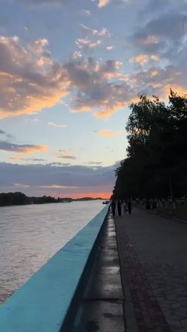A vertical video shows a river promenade at sunset. A wide, calm river is on the left, bordered by a light blue railing. A paved walkway with red and gray brick patterns runs along the right side of the river, lined with trees. Several people are walking on the promenade, casting shadows on the wet pavement. The sky above is a blend of blue, with scattered clouds tinged orange and pink by the setting sun. The horizon shows a distant, tree-lined shore.