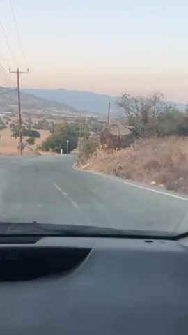 The video shows a view from inside a vehicle as it travels along a winding paved road in a rural, hilly area. The road has white lane markers and some cracks. There are power lines and poles on the left side of the road. On the right side of the road, there is dry brush, some trees, and a large boulder. In the distance, there are more hills and mountains under a clear sky with a soft, pastel hue, suggesting either dawn or dusk. There are no people visible in the video. A speed limit sign indicating 50 is visible on the left side of the road at one point.
