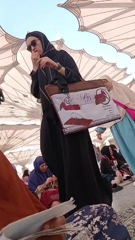 The video depicts a crowd of women dressed in traditional Islamic clothing moving under a large, white, tent-like structure, possibly outdoors. The women are wearing headscarves and long dresses. One woman carries a bag with a printed design and text in Arabic. Many of the women are either walking or seated on the ground. The ground is covered with various patterned rugs. The scene is brightly lit, suggesting it is daytime.