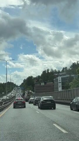 A daytime video shows a view from inside a vehicle driving on a multi-lane highway in Asker, Norway. The sky is partly cloudy with patches of blue. The road is experiencing heavy traffic, with numerous cars, including a large truck, moving slowly or at a standstill in multiple lanes. On the right side of the highway, there is a concrete barrier, beyond which are buildings nestled among green trees on a hillside. The buildings appear to be commercial or office structures. On the left side, the highway continues into the distance, also filled with traffic. Streetlights are visible along the median and sides of the road. Notable vehicles include a black Tesla Model S with license plate "ED 88418" in the foreground, and an Audi station wagon with license plate "SL 20043" in the left lane. Another dark SUV with license plate "EV 12382" is also visible in the right lane. The overall scene depicts a typical busy highway environment during the day.