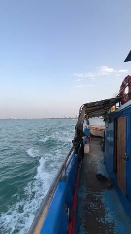 The video shows the view from the side of a blue boat moving through turquoise water. Waves lap against the hull, creating white foam. In the distance, a hazy city skyline can be seen. The deck of the boat is visible, with a weathered wooden door, a life preserver, and some items on the floor. The sky is a clear, pale blue.