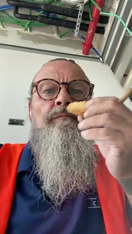 The media file is a close-up, head-and-shoulders shot of a man with a long, grey beard and round, tortoiseshell glasses. He is wearing a navy blue polo shirt and a bright orange safety vest. His mouth is open, and he is holding a biscuit or cookie to his mouth with his right hand. His expression is somewhat ambiguous, but he appears to be in the process of eating or about to eat the item.

The background suggests an indoor construction or renovation site. Exposed pipes, wiring, and ceiling beams are visible. The lighting is bright, indicating it is daytime. There are no clear indicators of the specific time of day beyond it being light. The location context provided is Ramot HaShavim, Israel.

The activity is simple: the man is eating. There are no other discernible events or interactions. Notable details include the man's distinctive beard and glasses, the safety vest suggesting he is on a work site, and the general context of construction. There is some faint text visible on the man's shirt, which appears to be a logo or brand name, but it is largely obscured and illegible. No other text is present in the media.