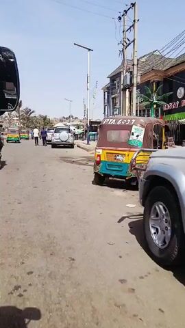 The video shows a street scene, filmed from a moving vehicle. The street has a worn asphalt surface with visible patches and marks. There are parked vehicles on the right side of the street, including a silver Toyota RAV4 with a spare tire on the back and a silver SUV, possibly a Toyota Highlander. Two men are walking along the street, near the parked vehicles. The man closer to the camera is wearing a white and light brown patterned short-sleeved shirt with dark pants, while the other man is wearing a blue short-sleeved shirt and dark pants. In the background, there are buildings, power lines, street lights, and other pedestrians. Some tricycle taxis, painted in yellow and green, are visible, as well. There are businesses along the side of the street, one marked "My Milles," and another that advertises "Ice Chill." A person in a pink outfit is near the "My Milles" storefront. The weather appears sunny and clear.